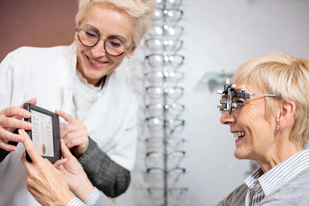 Smiling mature female ophthalmologist examining senior woman, holding an eye chart