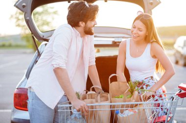 Bakkal alışverişdönen mutlu genç çift, bir araba bagajına gıda ile kağıt torba yükleme