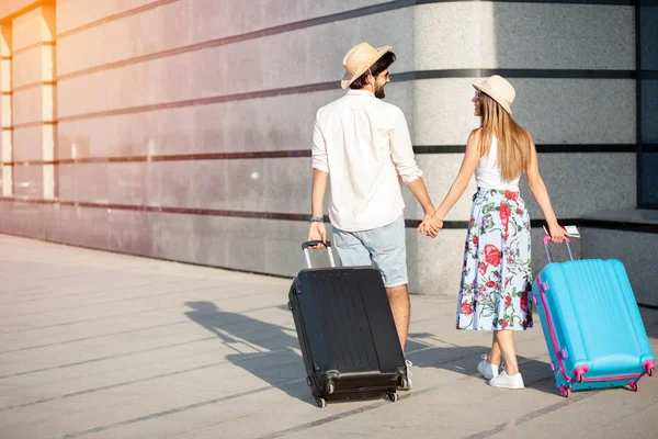 İki mutlu genç turist el ele kameradan uzaklaşıp bavullarını çekip birbirlerine bakıyorlar.