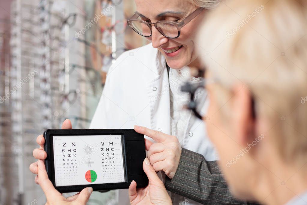 Smiling mature female ophthalmologist testing patient's vision for myopia with a test chart