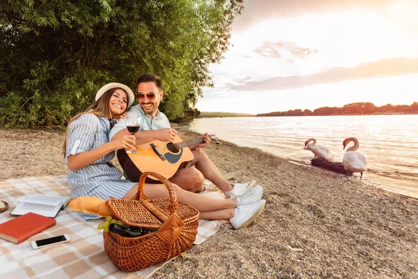El hombre está tocando la guitarra y su novia está apoyando su cabeza en su hombro. Puesta de sol sobre el agua en el fondo — Foto de Stock