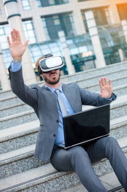 Genç işadamı VR gözlük kullanarak ve el jestleri yapma, bir ofis binasının önünde bir dizüstü bilgisayar üzerinde çalışıyor
