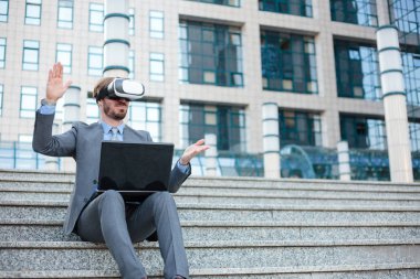 Genç işadamı VR gözlük kullanarak ve el jestleri yapma, bir ofis binasının önünde bir dizüstü bilgisayar üzerinde çalışıyor