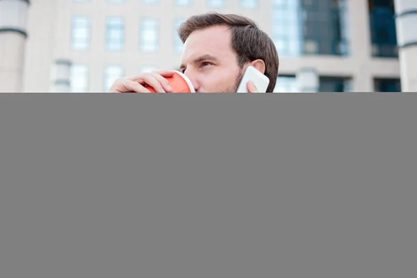 Νέος επιχειρηματίας που δουλεύει σε ένα φορητό υπολογιστή μπροστά από ένα κτίριο γραφείων, πίνοντας καφέ και μιλώντας στο τηλέφωνο. Κοντινό-up φωτογραφία — Φωτογραφία Αρχείου