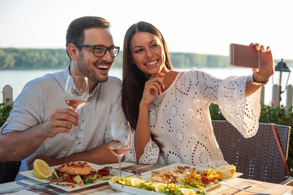 행복한 젊은 부부가 셀카를 찍기 위해 포즈를 취하고 강변 레스토랑에서 저녁 식사를 하고 미소짓고 있습니다. — 스톡 사진