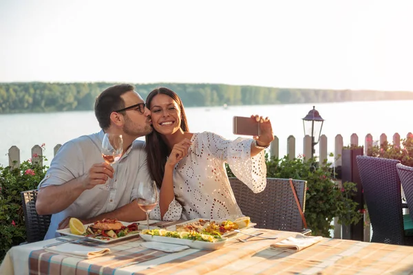 Romantik genç çift selfie için poz, nehir kenarındaki bir restoranda akşam yemeği yiyor