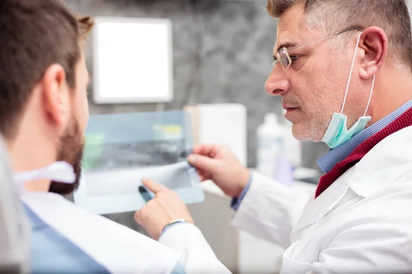 Odontólogo varón maduro que muestra una imagen de rayos X a un paciente varón joven durante el examen en la clínica dental —  Fotos de Stock