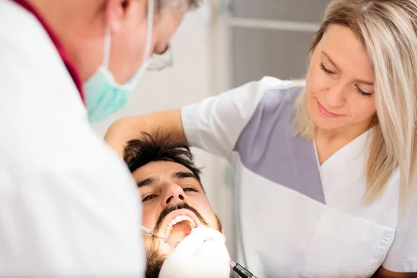 Dentista masculino maduro y hermosa asistente joven que realiza un examen dental en un paciente masculino joven en la clínica dental —  Fotos de Stock