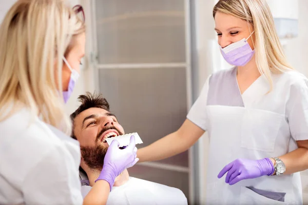 Dos dentistas jóvenes que trabajan en la clínica dental. Blanqueamiento de los dientes del paciente masculino y el uso de la tabla de tonos para mach color —  Fotos de Stock