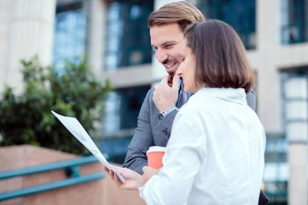 Two happy young business partners meeting in front of an office building, looking at sale charts and discussing project details