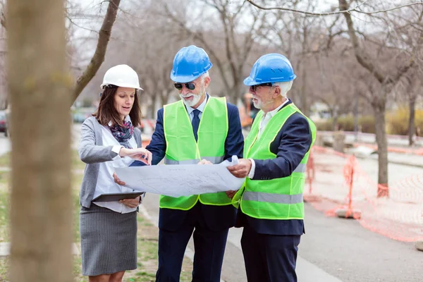 Equipo mixto de hombres mayores y jóvenes arquitectos o socios comerciales que buscan planes de construcción en un sitio de construcción — Foto de Stock