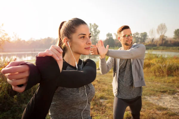 Determined young couple stretching arms during early morning workout by the lake or river, listening to music