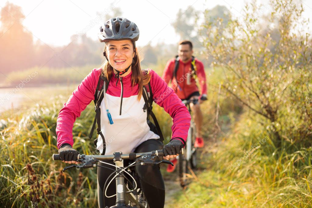 Beautiful happy young woman riding mountain bike over a meadow by the lake or river in the early morning