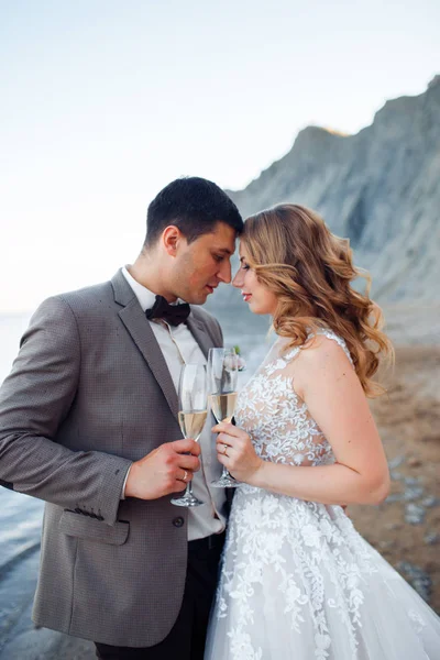 Bela noiva e noivo em um terno com champanhe em um contexto de montanhas . — Fotografia de Stock
