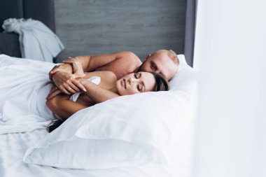 Sevgi dolu bir çift yatakta güler. Beyaz çarşaflar üzerinde birbirlerine gülümsüyorlar..