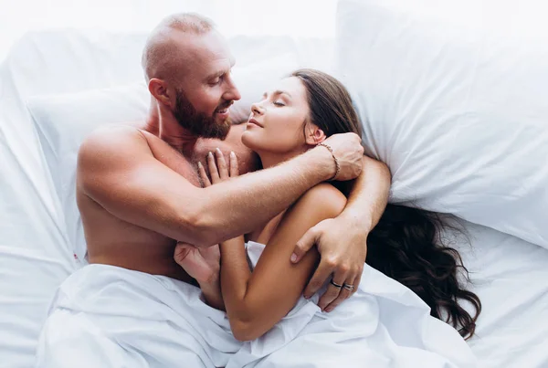 Um casal amoroso ri-se na cama. Sorrindo um para o outro em folhas brancas. — Fotografia de Stock