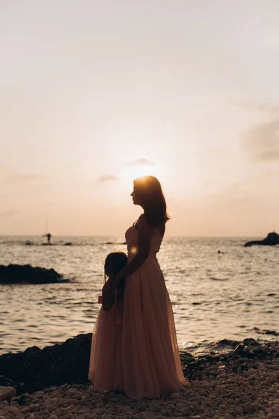 Mother and daughter on a cliff look at the sea in pink dresses on the Sunset.