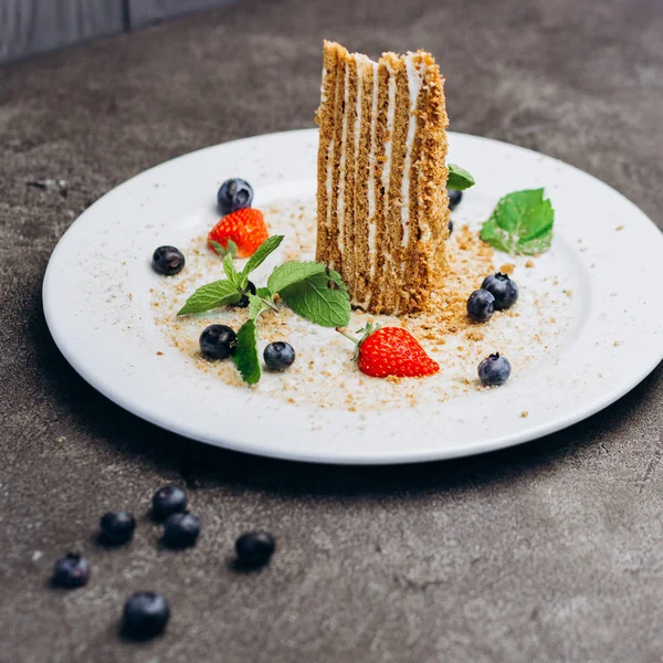 Slice of honey cake on plate decorated with berries on a gray background.