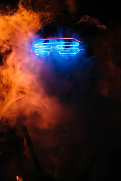 head in neon paint and glowing glasses in a hood a man close-up in neon paint smokes, face in neon and smoke