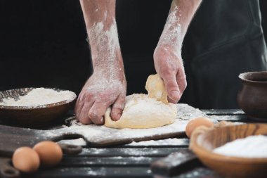 Güzel ve güçlü erkeklerin elleri hamuru yoğurarak ekmek, makarna ya da pizza yaparlar. Bir un bulutu toz gibi uçuşuyor. Tavuk yumurtasının yanında. Arka plan karanlık.