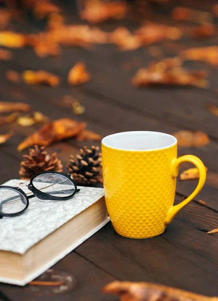 Yellow cup of tea with a book and glasses on a wooden background with a lot of dry fallen leaves. Relaxing scene. Selective focus. Bad weather.
