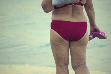 Kadın kalçaları yağ ile kaplıdır. Selülit. Mayo giyen kadın plajda kumda yürüyor. Mayo giymiş şişman kadın.