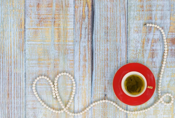 A small red porcelain cup with green tea on a saucer and a long string of white pearls on bluish vintage boards of a table with negative space