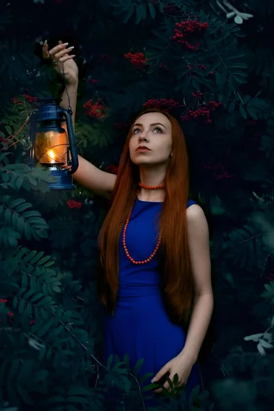 Portrait of a red-haired girl with an oil lamp and a face lit by fire among the branches of a mountain ash