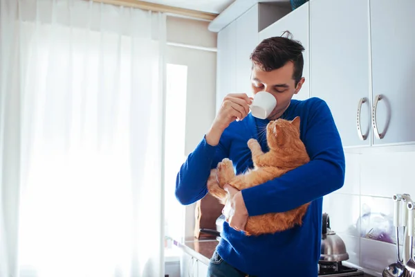 Yakışıklı genç adamın kedi tutan ve mutfakta çay içme portresi.