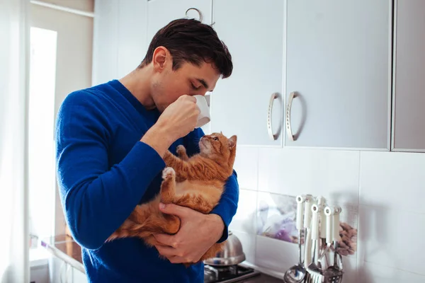 Yakışıklı genç adamın kedi tutan ve mutfakta çay içme portresi.
