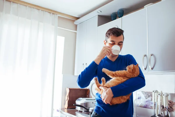 Yakışıklı genç adamın kedi tutan ve mutfakta çay içme portresi.