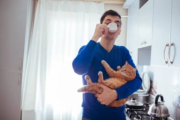Yakışıklı genç adamın kedi tutan ve mutfakta çay içme portresi.