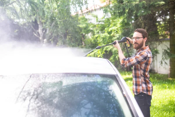 Car washing in the garden with a high pressure washer on a warm sunny day. A young bearded man using a compact home high pressure washer. Backyard, exterior, outdoor car cleaning.