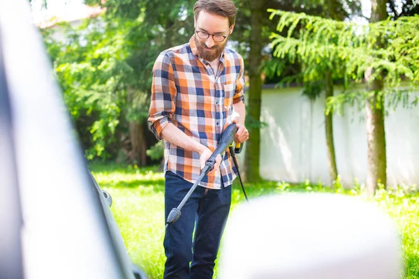 Car washing in the garden with a high pressure washer on a warm sunny day. A young bearded man using a compact home high pressure washer. Backyard, exterior, outdoor car cleaning.