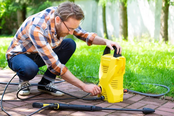 A young man works in the garden using a yellow compact home high pressure washer. Garden, patio, backyard cleaning on sunny day. An outdoor pressure washing, greenworks, cleaning car and exterior.