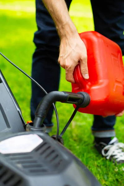 Man refilling a fuel tank in a petrol lawn mower with a red plastic canister with a funnel, close up. Gardening, greenworks, using a gasoline lawn mower, gasoline lawnmower in a garden on a sunny day