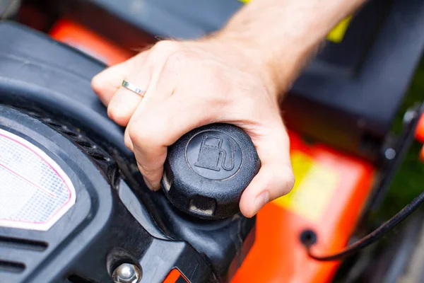 Refilling a fuel tank in a petrol lawn mower, close up on a hand on fuel filler cap. Male hand holding the fuel filler cap. Gardening, using a gasoline lawn mower in a garden on a sunny day