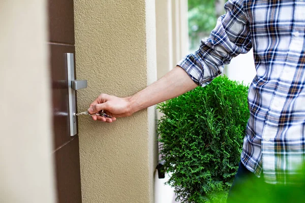 A man in casual clothes, opening, closing the front door of the house on a sunny day. Male hand holding a bunch of keys at an elegant house entrance. Focus on the hand inserting the key into the lock.