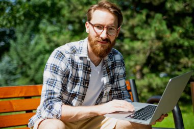 Parktaki bankta oturmuş, modern popüler ince dizüstü bilgisayarıyla kameraya bakan gündelik kıyafetli genç bir adam. Güneşli bir günde dışarıdaki bir bilgisayarda interneti kullanmak. Serbest çalışma..