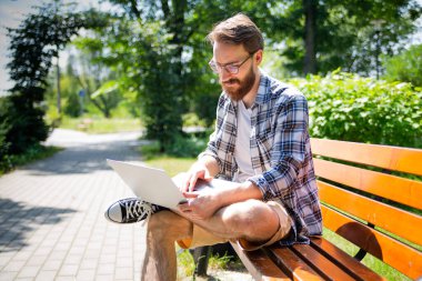 Sakallı ve gözlüklü genç adam gündelik giysiler içinde modern popüler ince dizüstü bilgisayarıyla bir bankta oturuyor. Güneşli bir günde dışarıdaki bir bilgisayarda interneti kullanmak. Serbest çalışma..