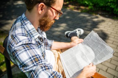 Gözlüklü genç sakallı bir adam, günlük kıyafetler giyiyor, bir bankta oturuyor ve günlük gazete okuyor. Güneşli bir günde açık havada rahatlamak.