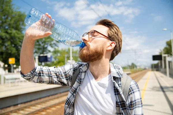 Tren istasyonunda bir platformda duran bir turist şişeden su içiyor. Genç yetişkin bir adam, sırt çantalı bir gezgin Polonya, Avrupa 'da tren beklerken içme suyu içiyor..
