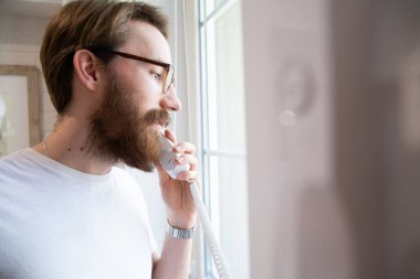 Beyaz tişörtlü, gözlüklü, elinde ev telefonu ahizesi olan parlak bir iç mekânda camdan dışarı bakan sakallı genç bir adam. Giriş telefonuyla kapıyı açmak için ahizeyi kaldırıyorum..