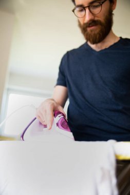 Adam ütü masasındaki kıyafetleri modern elektrikli buhar ütüsüyle ütülüyor. Günlük ev işleri, angarya, parlak bir iç mekânda ütü, arka planda bulanık bir adama odaklan.. 