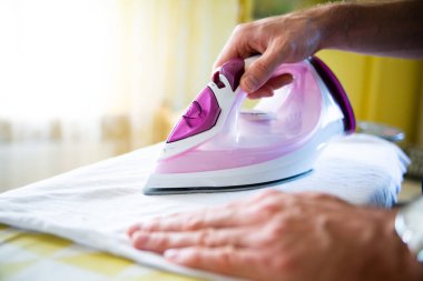 Erkek eli ütü masasındaki kıyafetleri modern elektrikli buhar ütüsüyle yakın çekimde ütülüyor. Ev işleri, angarya, parlak bir iç mekânda ütü, bir demire odaklanma.. 