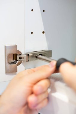 Beyaz mutfak dolabının kapağındaki menteşeleri parlak bir iç mekânda tornavidayla onarıp onarıyor. Tamircinin alet edevat, odaklanma ve menteşeye yakın çekim ile yaptığı küçük ev onarımı..