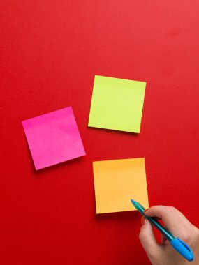 Üç renkli hatırlatma etiketinin üst görüntüsü ve kırmızı arkaplanda mavi bir kalem tutan bir kadın eli, metin için yer var.