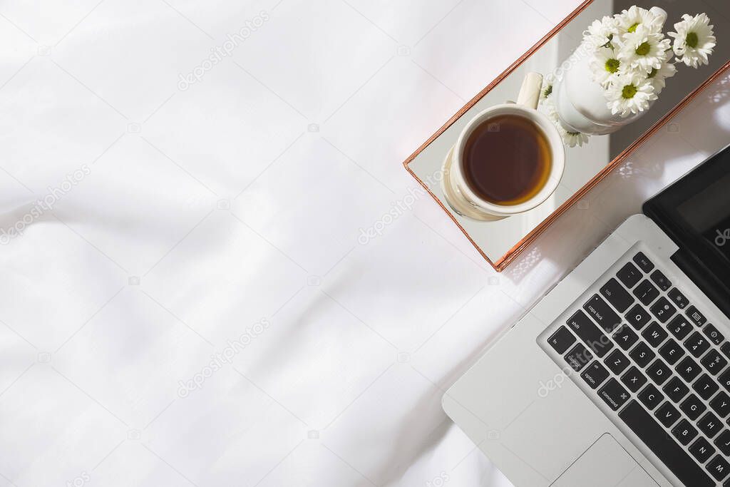 Top view of morning scene in voile fabric background with a laptop, mug of tea and a vase of white flowers in a mirrored brass tray, with space for text