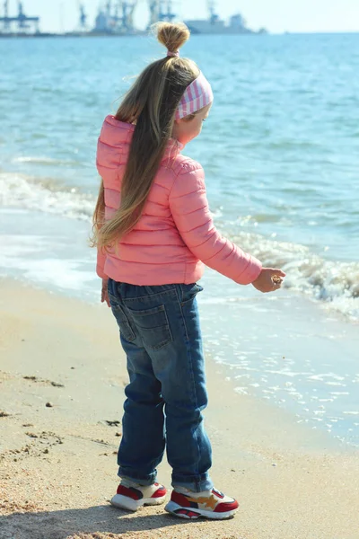 Una bambina con lunghi capelli biondi gioca sulla spiaggia con sabbia e conchiglie e guarda le onde del mare in una giornata di sole . — Foto Stock