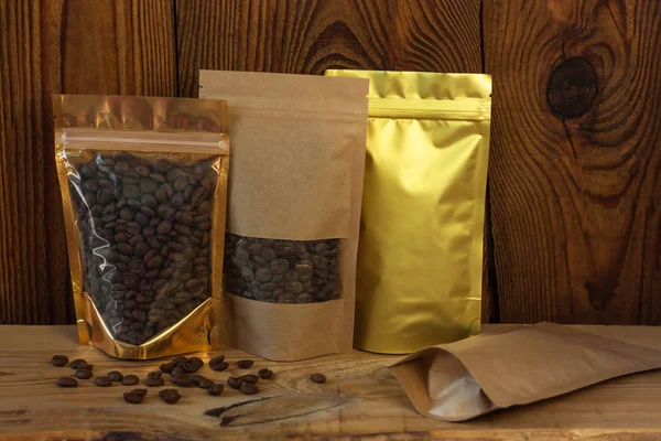 Papel kraft marrom e sacos de bolsa metalizados dourados com grãos de café vista frontal em um fundo de madeira . — Fotografia de Stock